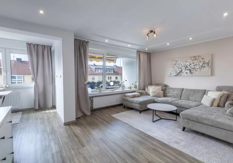 Modernes Wohnzimmer mit grauem Ecksofa, Holzboden und großen Fenstern mit neutralen Vorhängen.