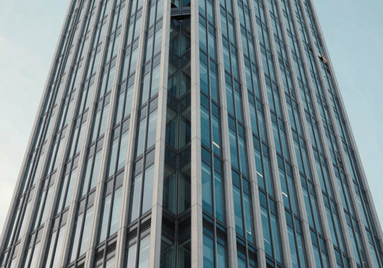 An abstract, low-angle photograph of a modern skyscraper's steel and glass facade against a light blue sky. The composition conveys strength, stability, and a future-forward vision. Muted blue and off-white color tones.
