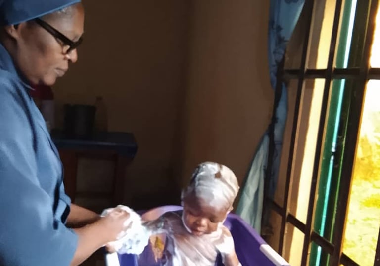 A caregiver bathing a small child with soap in a purple plastic tub indoors near a window.
