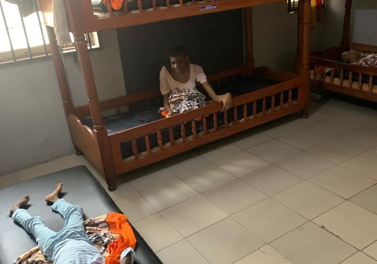 A person sitting on a wooden bunk bed and a child resting on a floor mattress in a shared dormitory room.