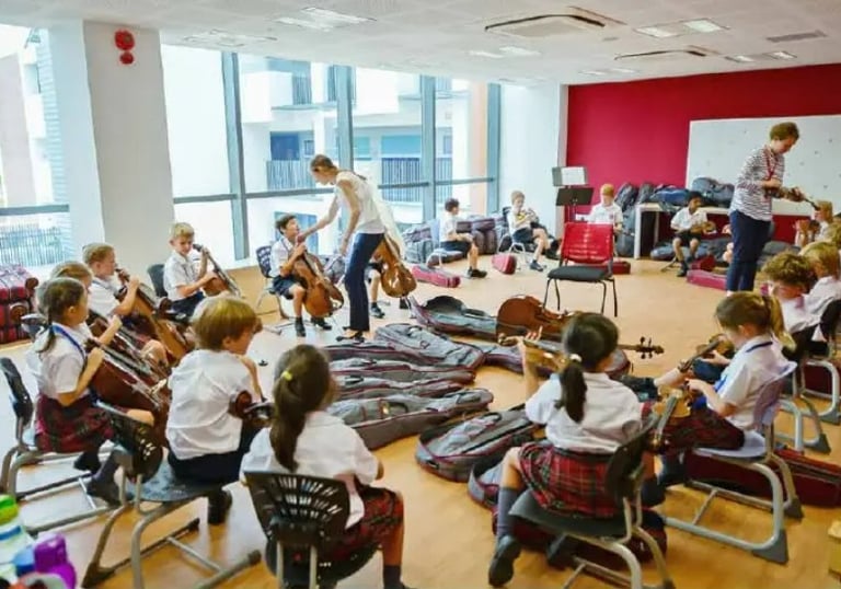 Students in school uniforms practice playing cellos and violins during a music class in a bright classroom.