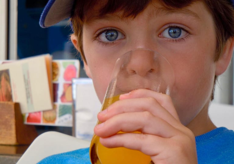 A young boy with striking blue eyes drinks a glass of orange juice at an outdoor cafe.
