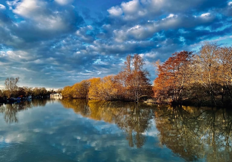 Tranquil river landscape with autumn trees reflecting in calm water under a dramatic blue cloudy sky.
