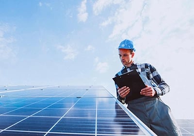 A solar technician in a hard hat inspects a residential photovoltaic panel system with a clipboard.