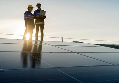 Two solar technicians in hard hats and safety gear inspecting rooftop solar panels with a laptop.