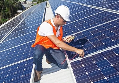 A solar panel installer in a safety vest and hard hat testing blue photovoltaic panels on a roof.