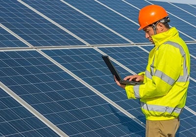 Solar energy technician in a hard hat using a laptop to inspect a solar panel farm.