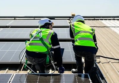 Two technicians in safety gear and hard hats installing solar panels on a rooftop for renewable energy.