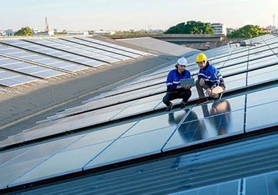 Two solar technicians in hard hats inspect a rooftop photovoltaic panel system with a laptop.