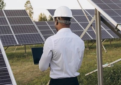 Solar energy engineer in a white hard hat using a laptop to inspect a solar panel field.