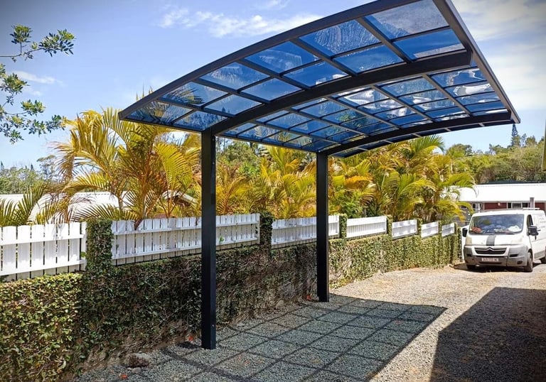 Modern black metal carport with blue tinted glass panels over a gravel driveway next to palm trees.