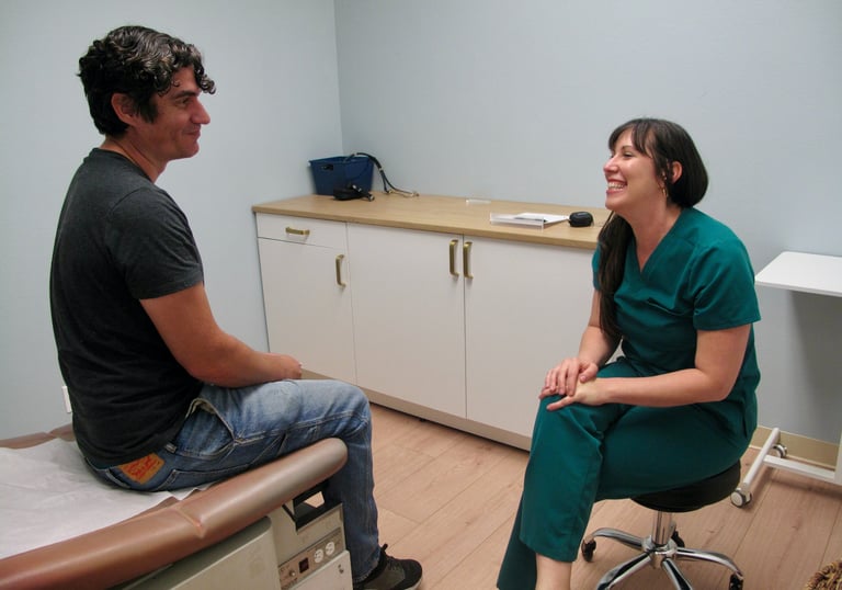 A smiling doctor in green scrubs consulting with a male patient in a medical exam room.