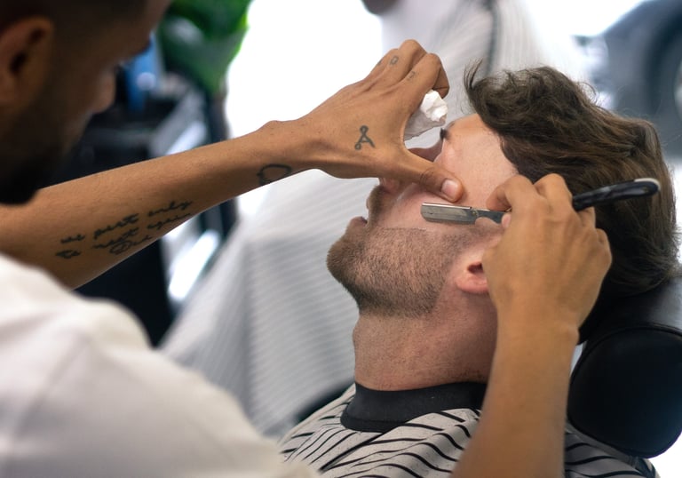 man receiving a shave with a cut throat razor by a barber while sitting in a chain in a barbershop