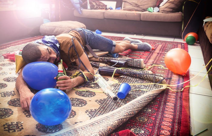 A man sleeping on a rug in a messy room after a party with balloons, beer bottles, and streamers.