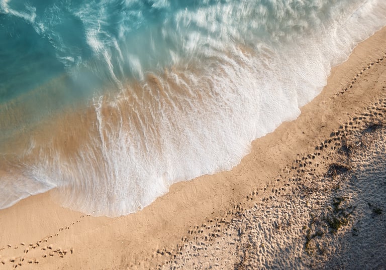 Aerial view of foamy turquoise waves crashing onto sandy beach with footprints along the shore.