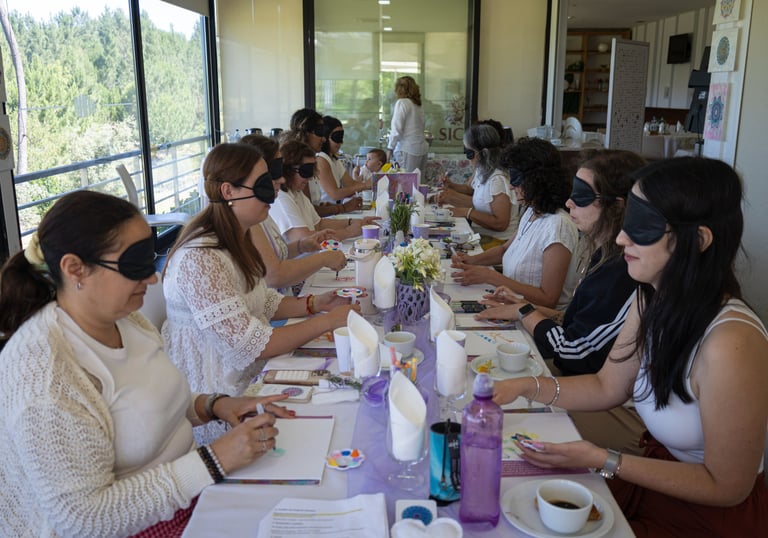 A group of women wearing blindfolds participating in a sensory art workshop at a long table.