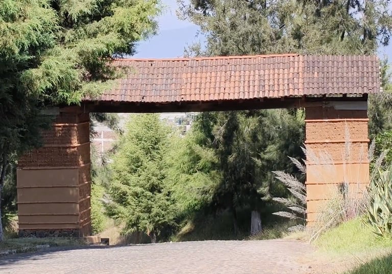 Patzcuaro Rustic stone archway with a clay tile roof over a paved road surrounded by lush trees.