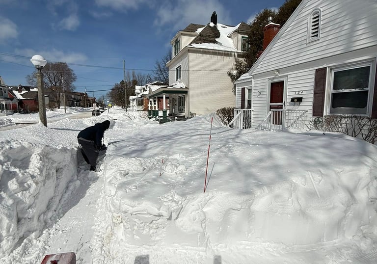 shoveling a deep snow drift on a sidewalk near a snowblower.