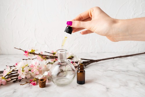 Hand using a glass dropper to put organic essential oil into a bottle near pink cherry blossoms.