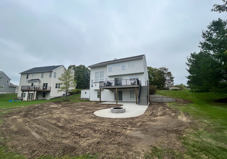 Backyard landscaping project with a new concrete patio, fire pit, and wooden deck under a cloudy sky.