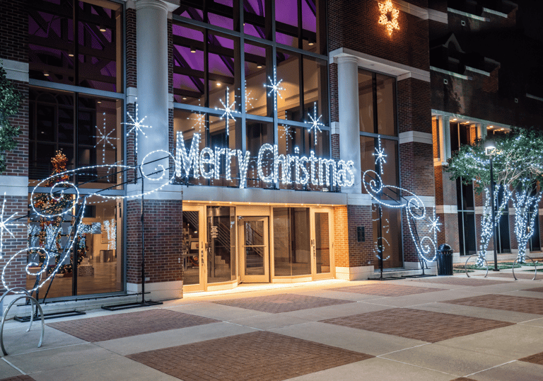 A brick building entrance decorated with a glowing white Merry Christmas neon sign and festive holiday lights.