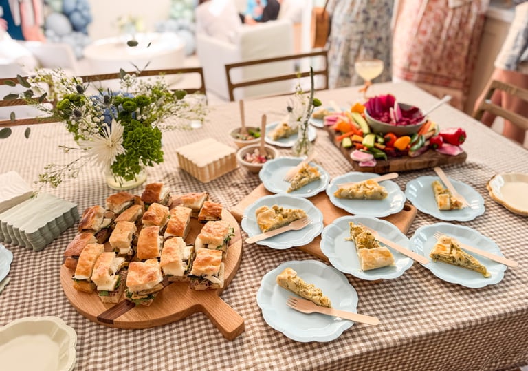 A catered party table with mini sandwiches, savory quiche slices, and fresh vegetable crudité.