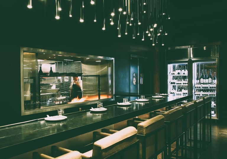 Modern upscale restaurant interior featuring an open kitchen, bar seating, and hanging pendant lights.