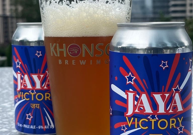 A glass of foamy Khonso Brewing pale ale next to Jaya Victory beer cans on a rooftop.