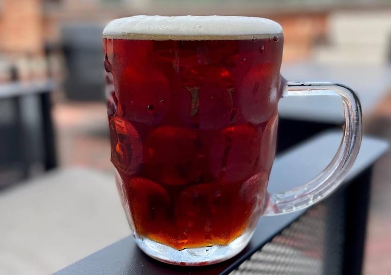 A cold dimpled glass mug of mexican dark amber ale with white foam sitting on a patio railing.
