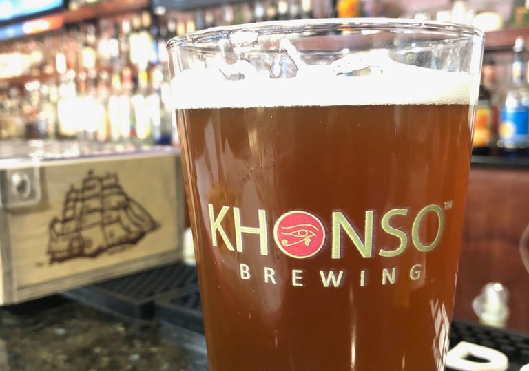 A pint of amber craft beer from Khonso Brewing sits on a bar counter with a blurred tavern background.