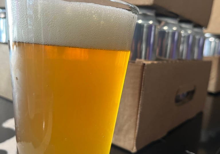 A cold pint of golden craft beer with foam head next to cardboard cases of canned beverages.