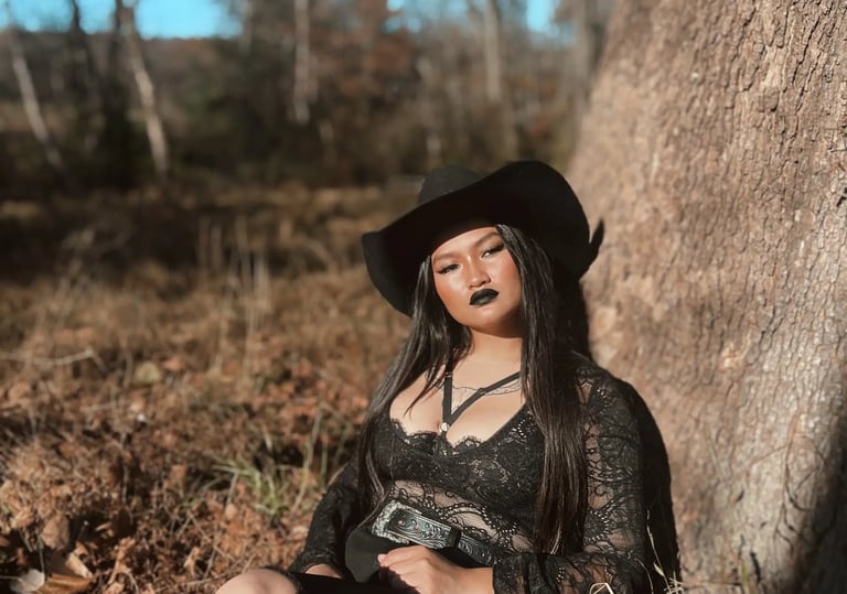 Amelia leaning against a tree in a black lace top with a black cowboy hat