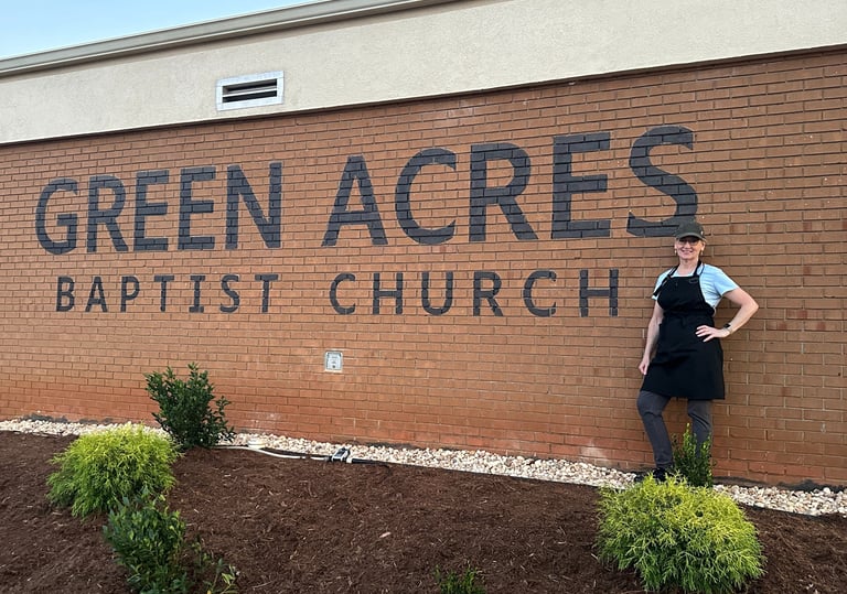 A smiling woman in an apron stands before the Green Acres Baptist Church brick sign.