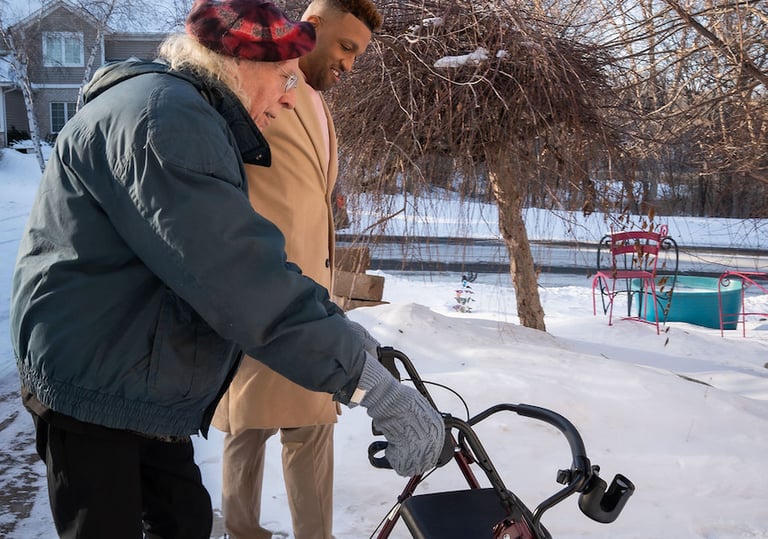 A caregiver assists a senior man using a walker on a snowy sidewalk during winter.