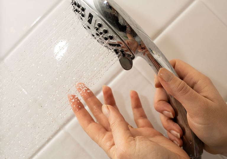 Hand testing the water temperature from a chrome handheld shower head against white tiles.