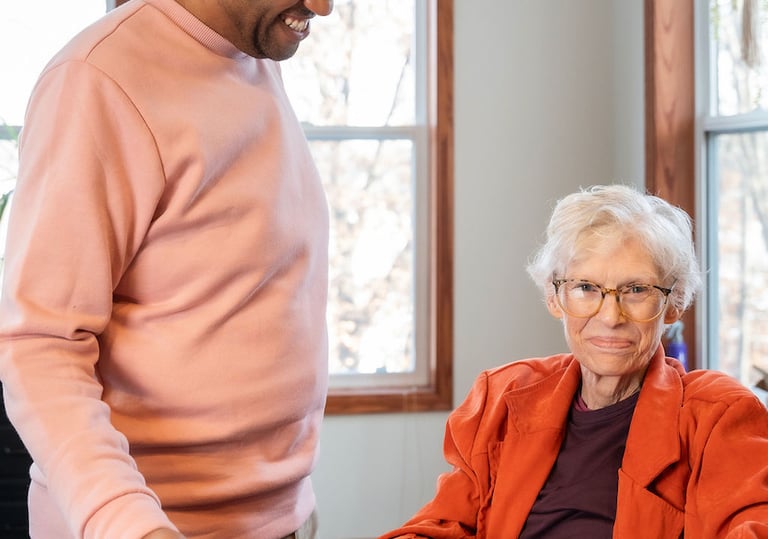 A smiling male caregiver serves a healthy meal of chicken and broccoli to a senior woman at home.