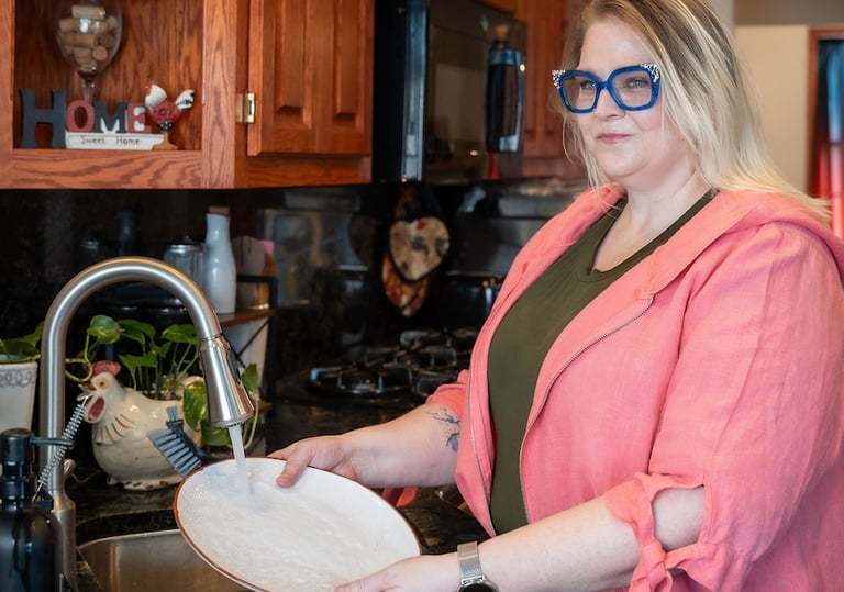 A blonde woman in a pink jacket washes a white plate in a stainless steel kitchen sink.