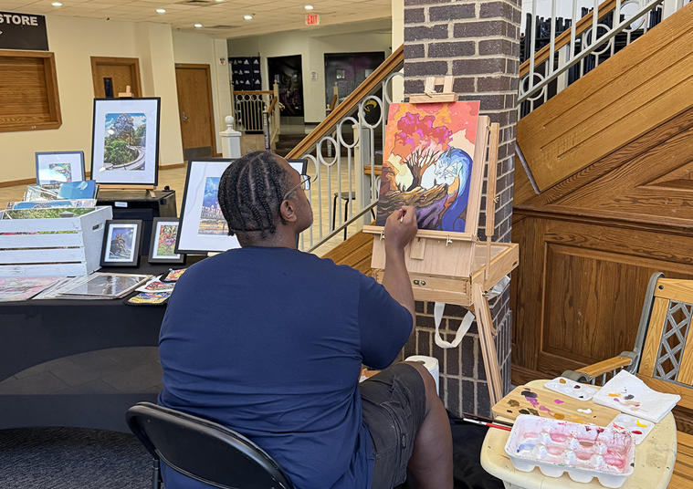 An artist with braided hair painting a colorful landscape canvas on an easel at an indoor art gallery.