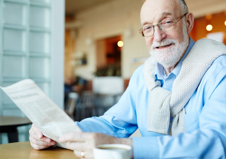 Senhor idoso, de barba, sorridente, de camisa social azul e blusão nas costas lê jornal.