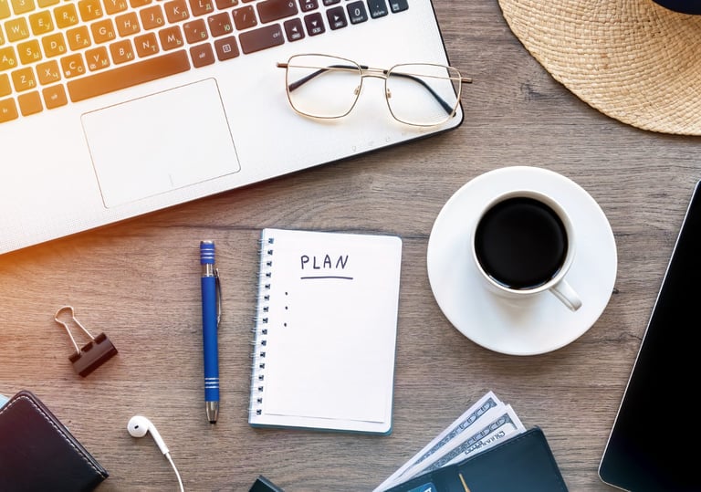 Uma mesa com notebook, xícara de café, caderneta, óculos, lapiseira, fone de ouvido e brochura.