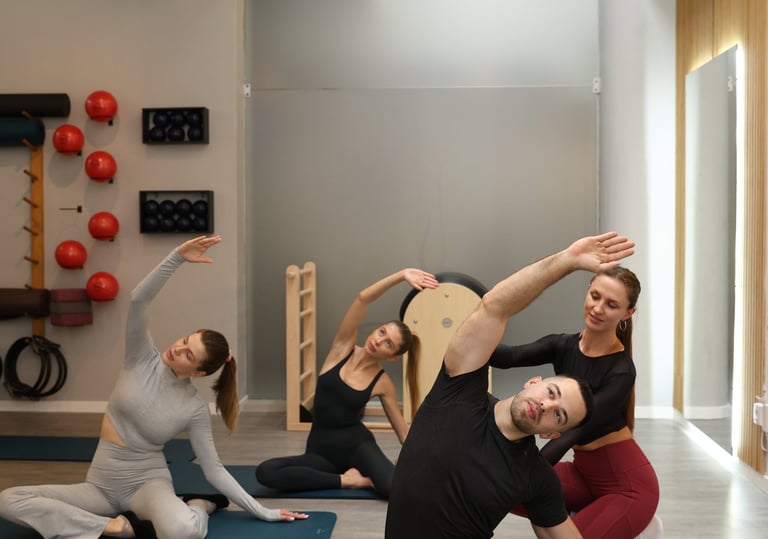 Aula de grupo de Pilates a realizar um alongamento lateral sentado nos tapetes, com uma instrutora