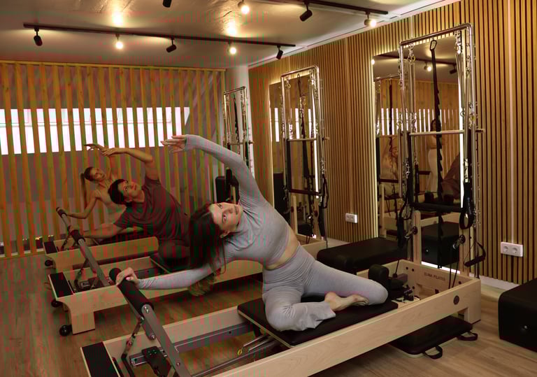 Três pessoas a fazer um alongamento lateral em reformers num estúdio de Pilates com espelhos 