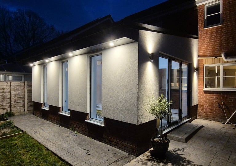 Modern home extension at night with bright soffit spotlights and outdoor wall lighting on white render.