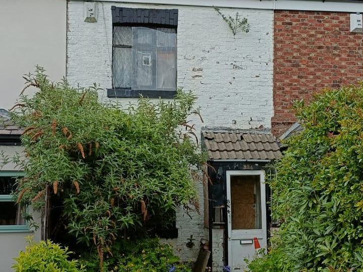 Abandoned white brick terraced house with overgrown garden and boarded windows under a blue sky.