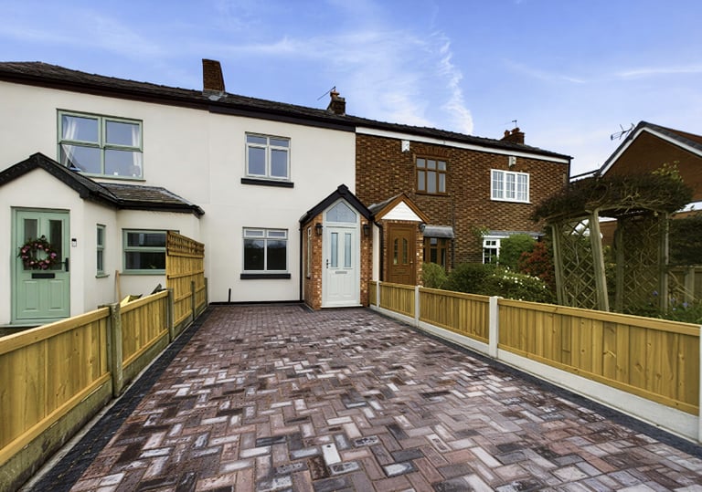 A charming terraced house with a red brick driveway and light wooden fences, featuring green and brown doors under a blue