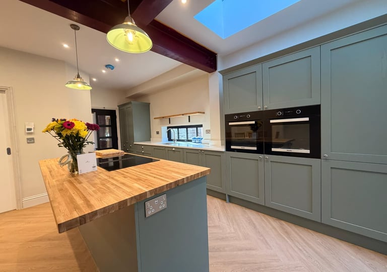 Modern sage green shaker kitchen with wooden butcher block island and integrated double ovens.