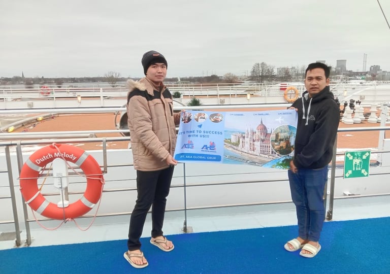 Two crewmembers hold an AKA Global Grup banner on the deck of the Dutch Melody river cruise ship.