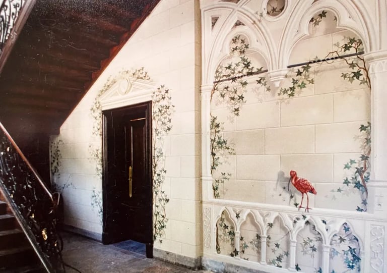 Hallway with wooden staircase and large mural of faux sandstone, ivy, gothic arches, and red bird.