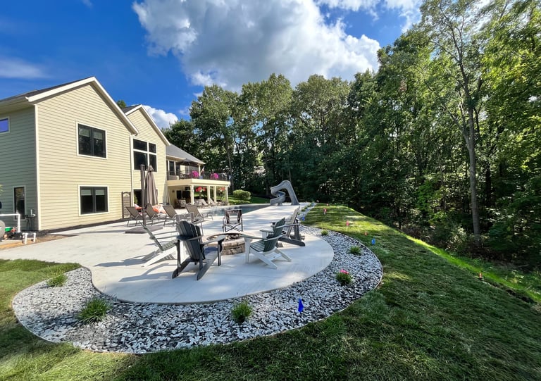 Modern backyard concrete patio with a fire pit, Adirondack chairs, and a swimming pool slide near lush green trees.