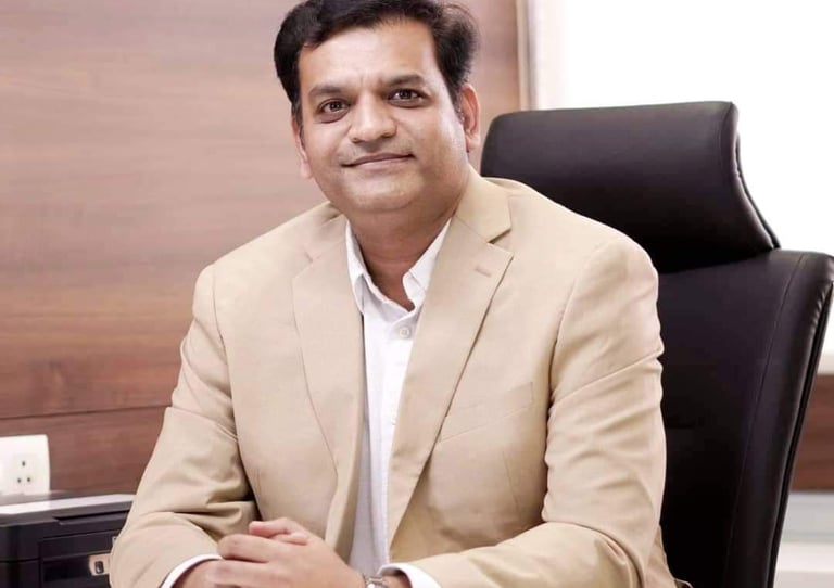 Professional portrait of a male business executive in a beige blazer sitting at an office desk.